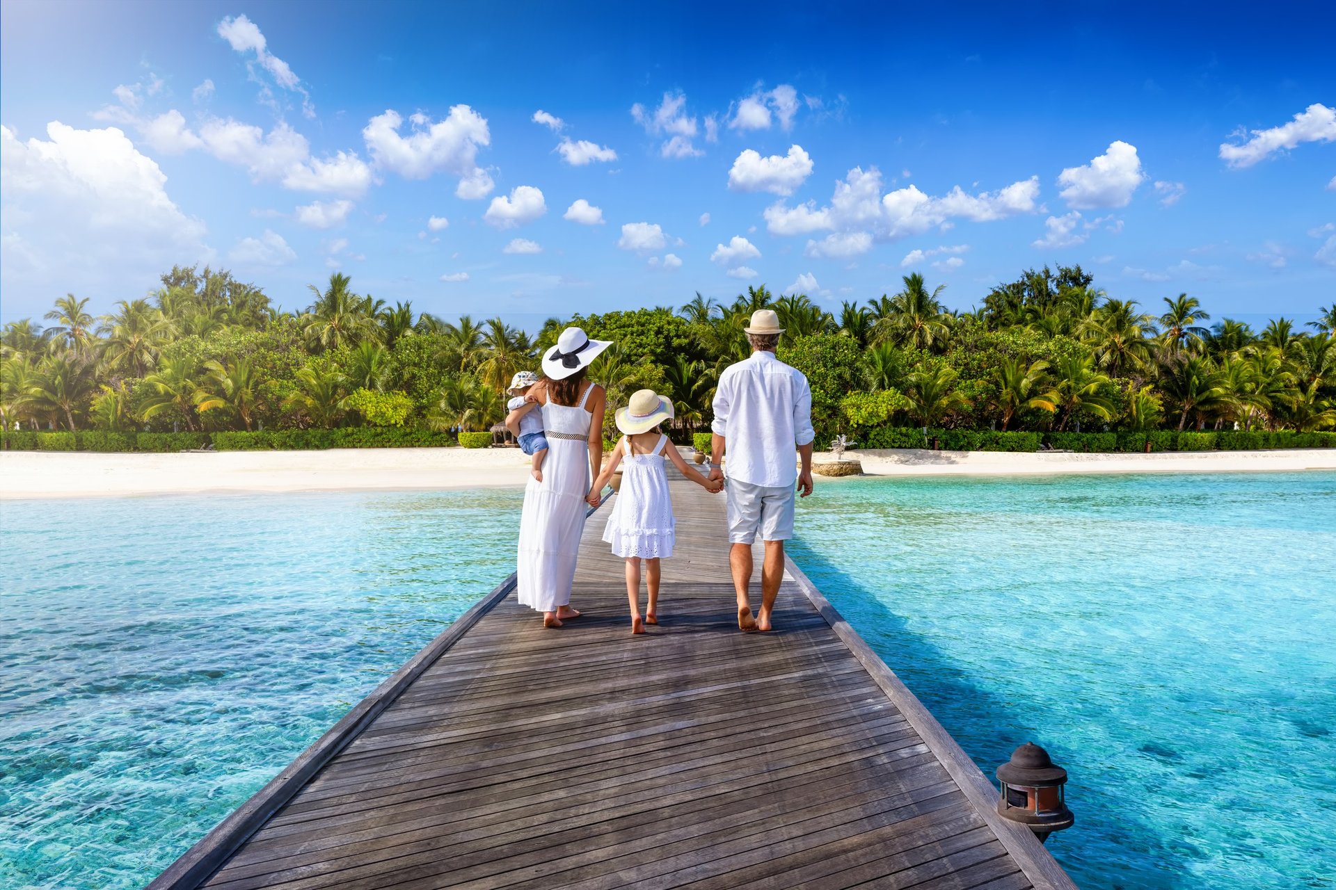 A family in white clothing walks on a pier towards a tropical island paradise with turquoise ocean and palm trees