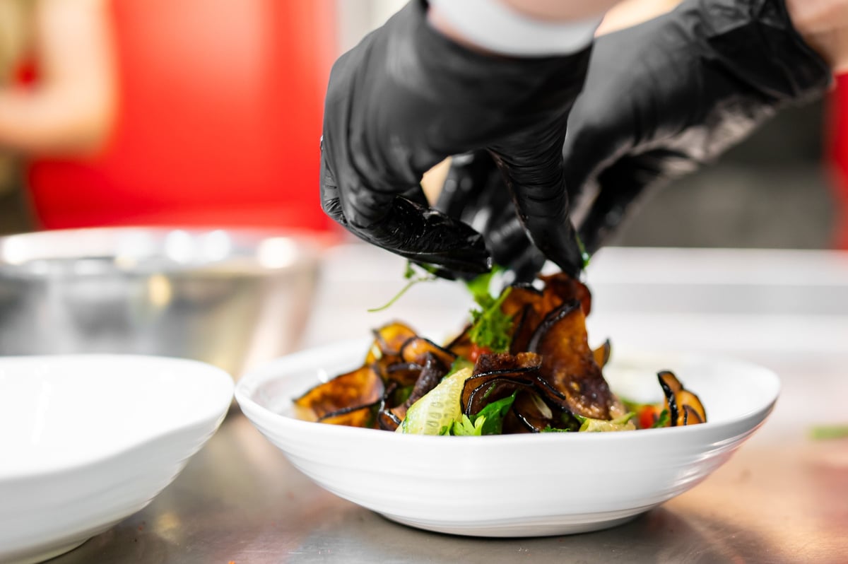 A chef wearing black gloves carefully garnishes a fresh dish in a commercial kitchen