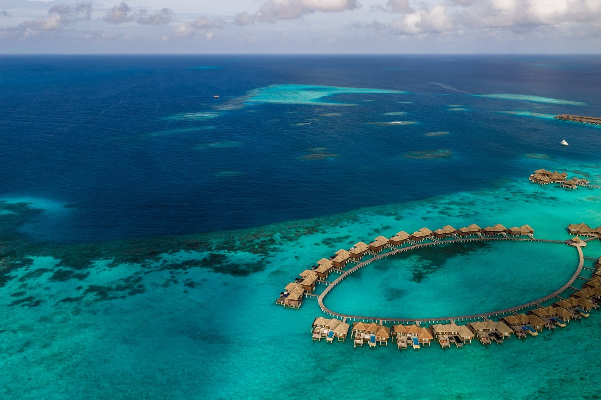 Aerial Maldives resort shows ring shaped overwater villas, circular boardwalk, thatched bungalows with decks and pools in turquoise lagoons
