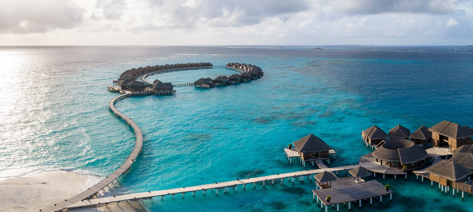 Aerial scene in Maldives shows a curved boardwalk to a ring of water villas above turquoise shallows, stilted bungalows with decks and plunge pools