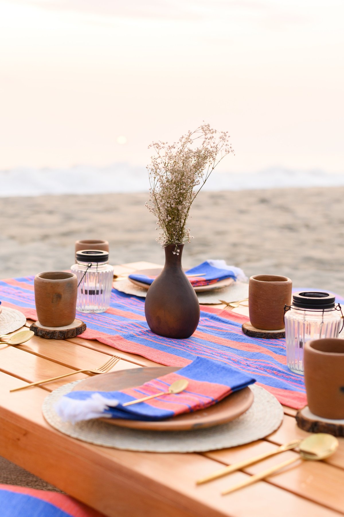 Bohemian table setup featuring natural plates, colorful napkins, golden cutlery, and dried flowers, creating a romantic outdoor dining experience on a beach at sunset