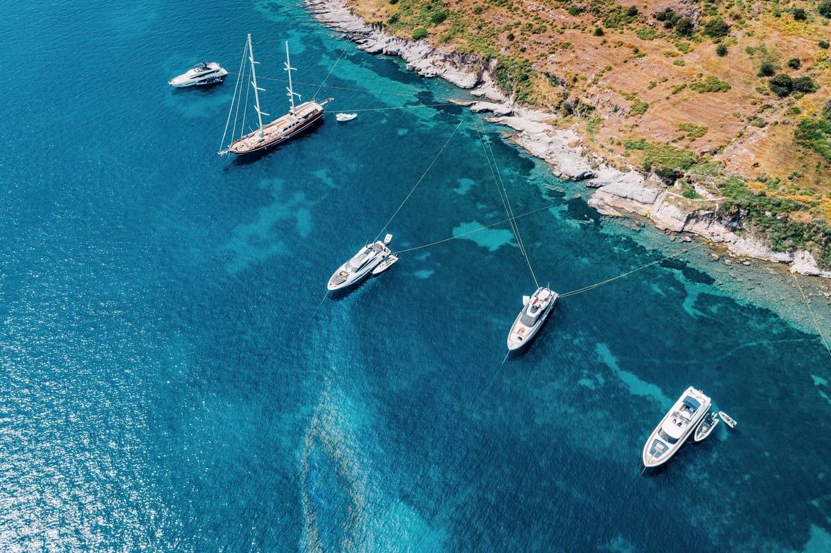 Aerial view showing several luxury yachts and sailing boats anchoring near a rocky coastline with clear turquoise blue water
