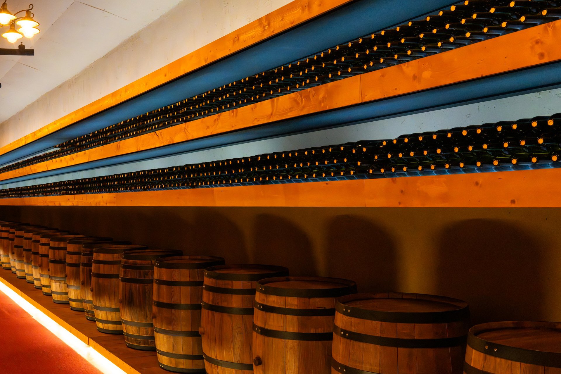 A wide-angle horizontal shot showing a dramatic perspective of long shelves filled with wine bottles above a row of wooden wine barrels along a lighted walkway in a wine cellar