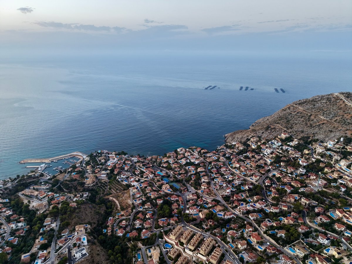 High altitude aerial view shows coastal town packed with villas, marina, swimming pools, winding roads and rocky cliffs beside calm blue sea