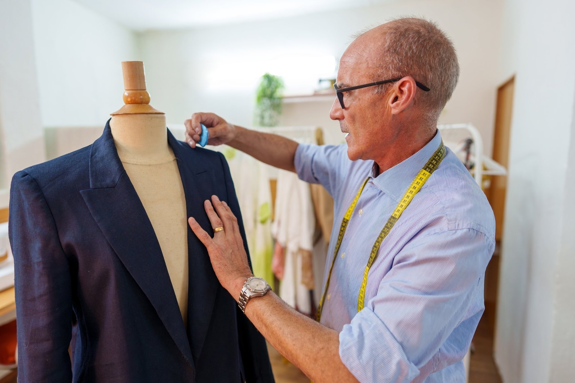 Mature tailor with measuring tape marking a suit jacket on a mannequin, working in a professional workshop