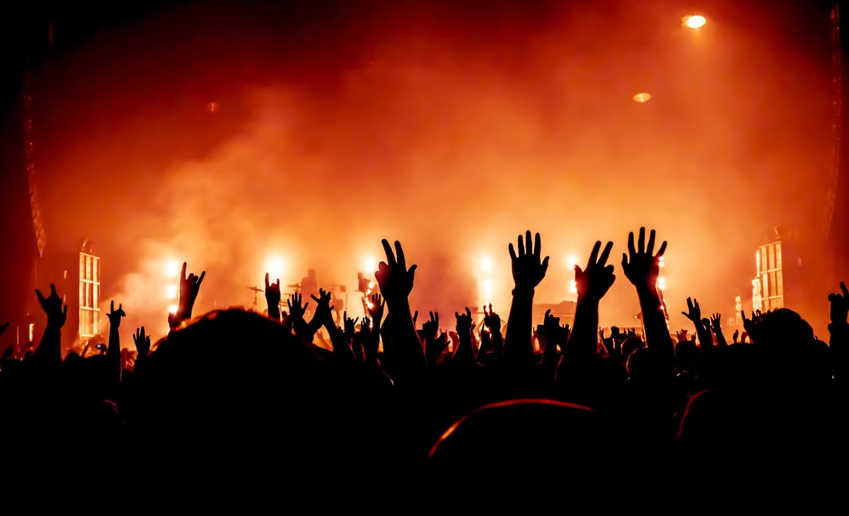 Silhouettes of a crowd with raised hands at a concert, with bright stage lights and orange fog in the background