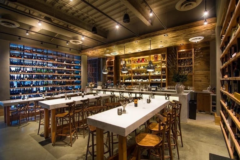 Modern bar interior with white dining table, wooden bar stools, and illuminated shelves displaying bottles of liquor and spirits