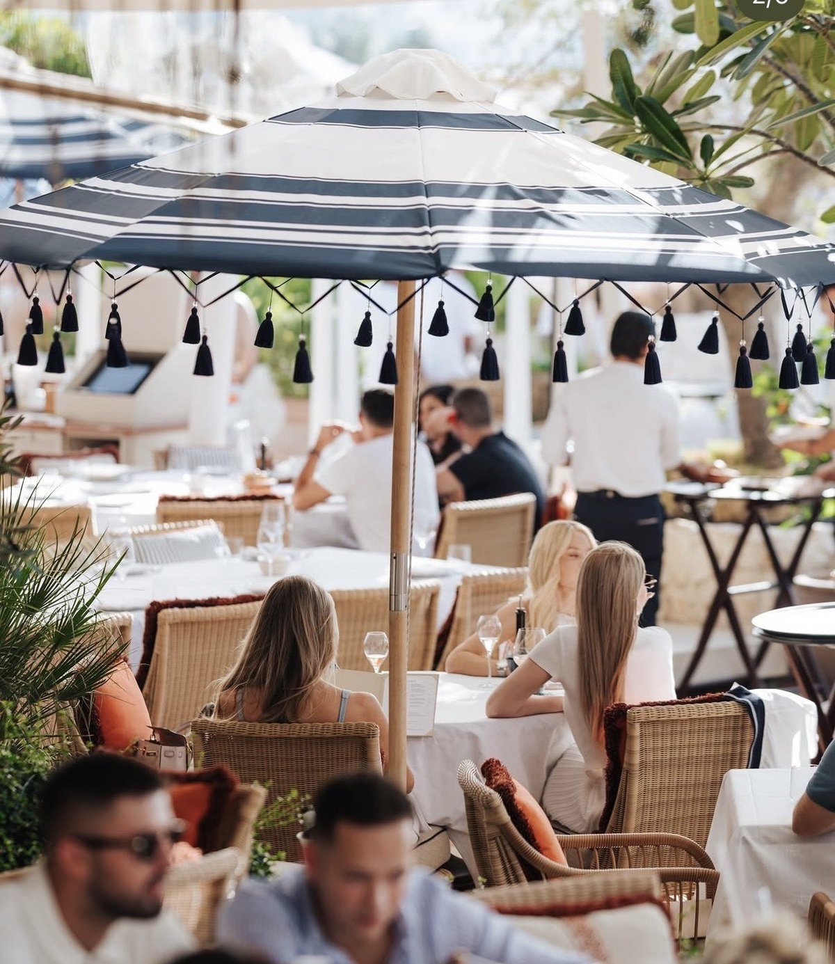 Outdoor restaurant patio with striped umbrellas, diners seated at tables with white tablecloths, and potted plants in natural daylight