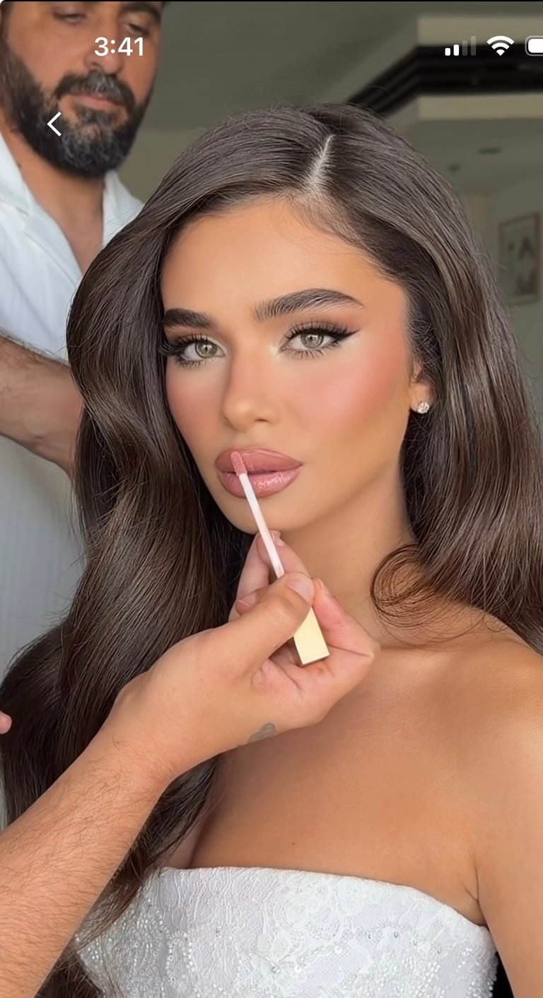 Woman having makeup applied by a man, holding a lip gloss applicator, with long brown hair and bare shoulder visible