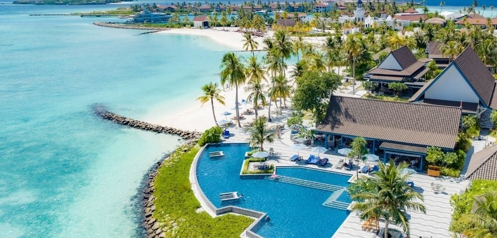 Aerial view of a tropical resort with curved blue pool, beachfront villas, palm trees, and turquoise ocean waters