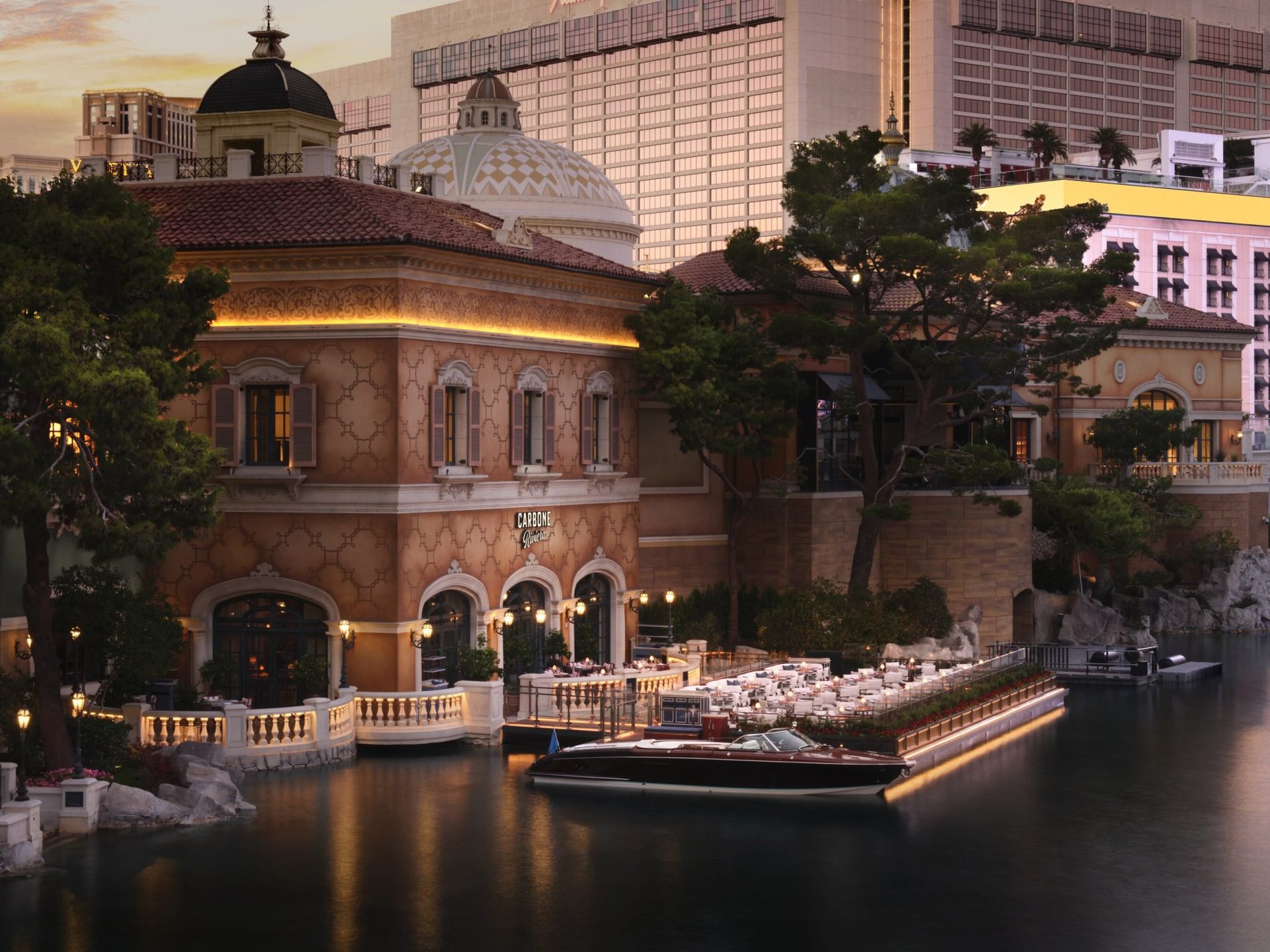 Illuminated historic brick building with dome beside waterway at dusk, surrounded by modern skyscrapers and trees
