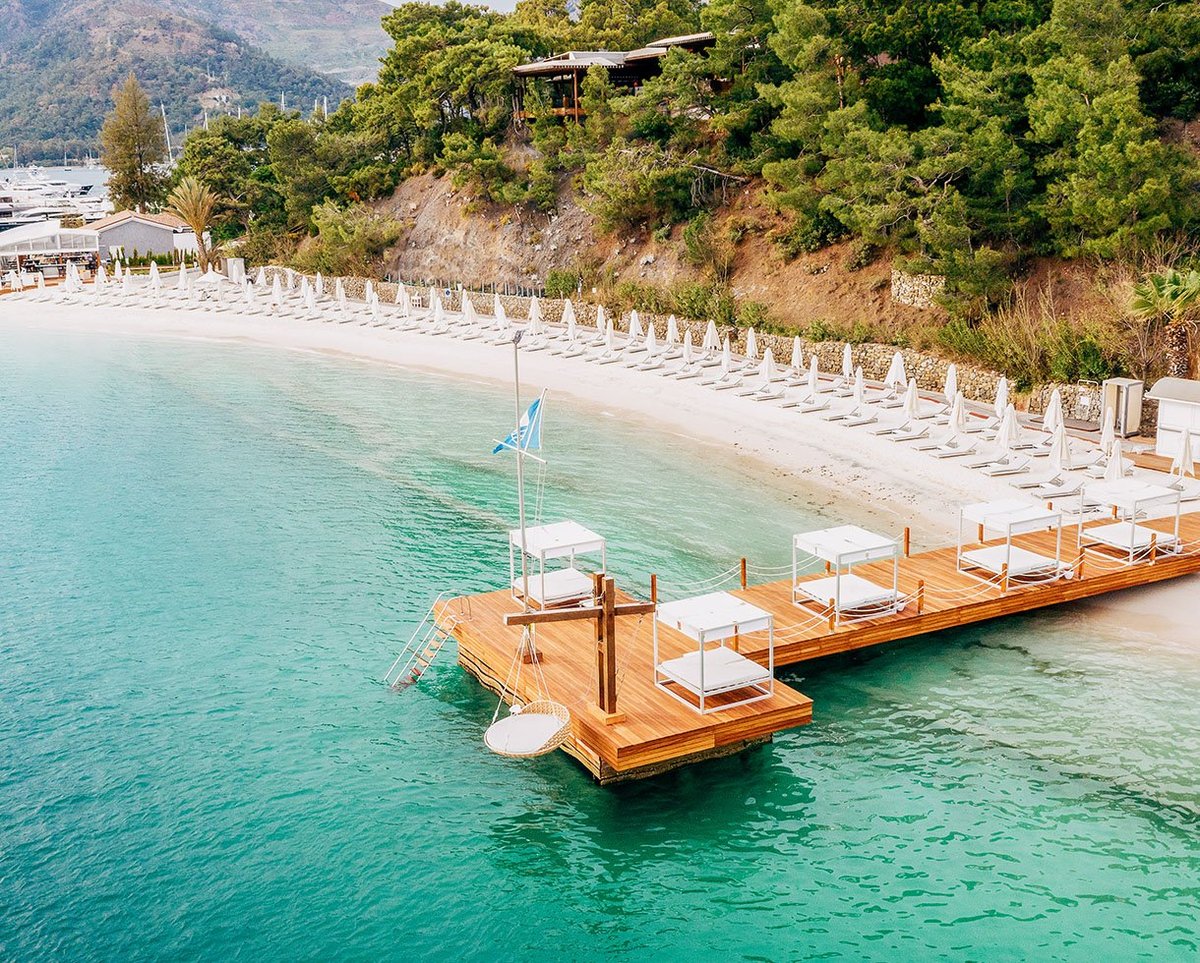 Luxury beach resort with wooden dock, sailboat, turquoise water, white umbrellas, and forested hillside