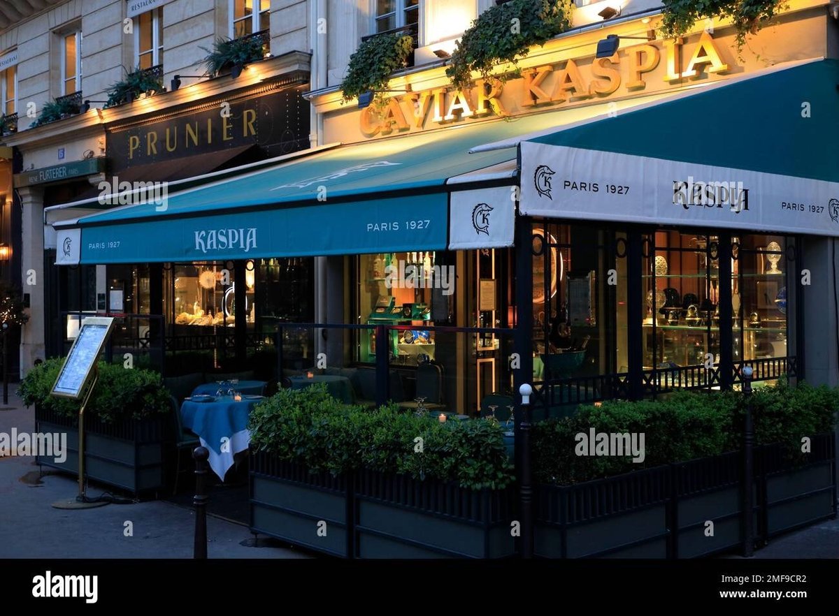 Parisian storefront with blue awning and illuminated signage at dusk, featuring shop windows and potted plants