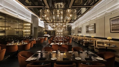 Elegant upscale restaurant dining room with rows of tables set with place settings, brown leather chairs, chandeliers, and modern lighting