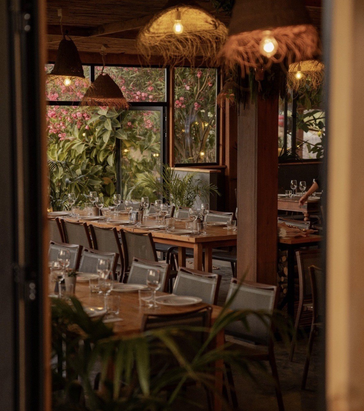 Elegant restaurant dining room with wooden table, gray chairs, large windows overlooking garden, woven pendant lights, and place settings