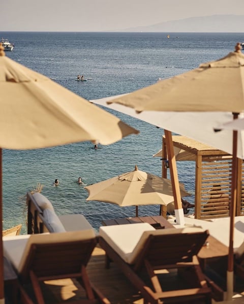 Beach lounge area with tan umbrellas, wooden loungers, and turquoise sea with swimmers in background