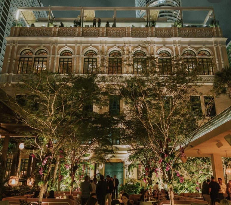 Historic ornate building at night with illuminated arched windows and decorated potted trees in foreground courtyard with visitors