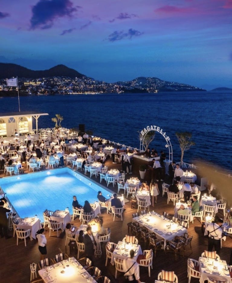 Luxurious beachfront wedding venue at dusk with illuminated pool, dining tables, and coastal mountains in background