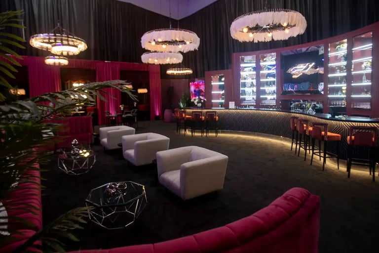 Modern lounge interior with pink and black decor, curved seating, illuminated pendant lights, and lit display cases along the back wall