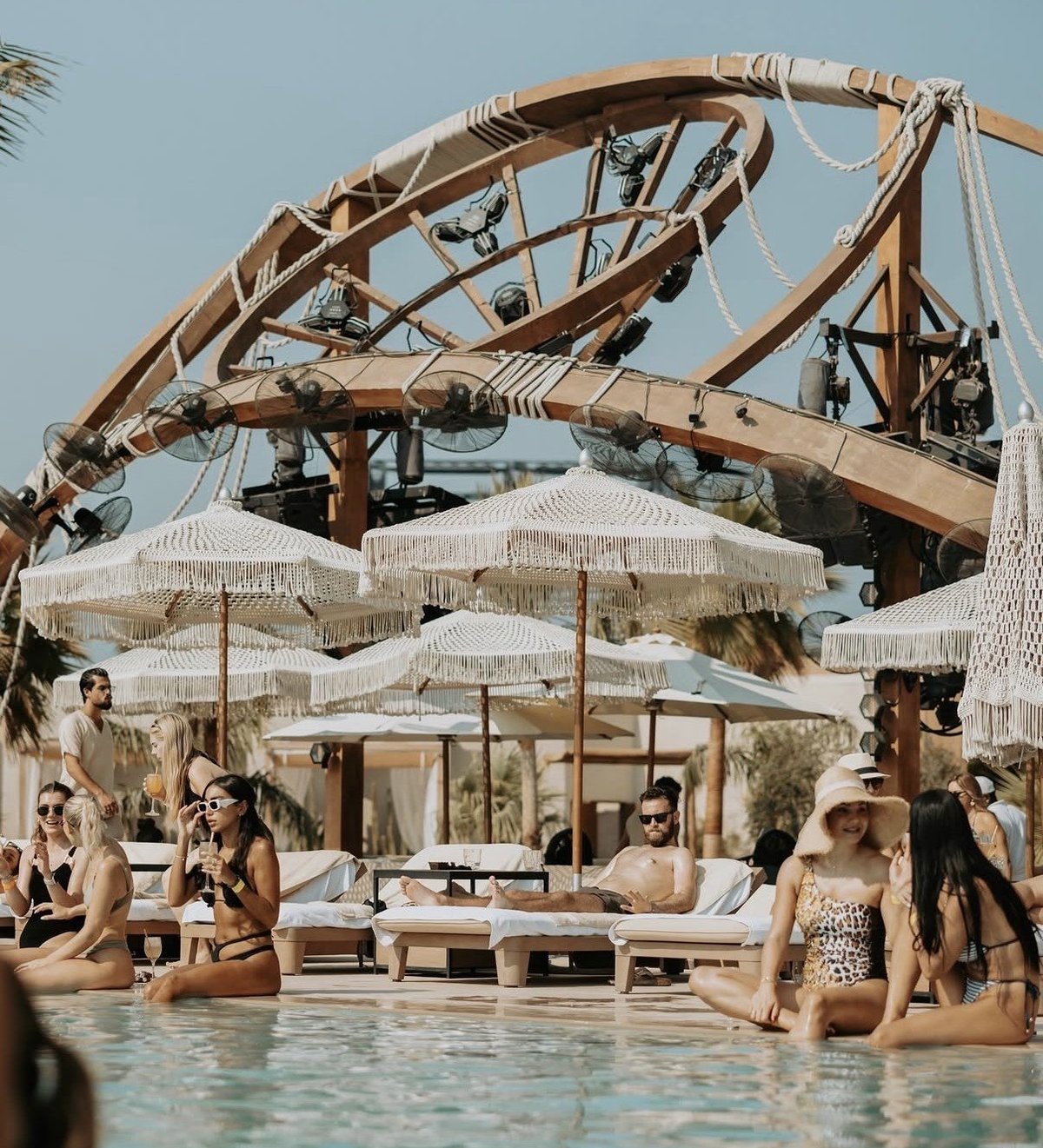 Luxury pool resort with wooden pergola structure, white umbrellas, and visitors relaxing in the water on a sunny day