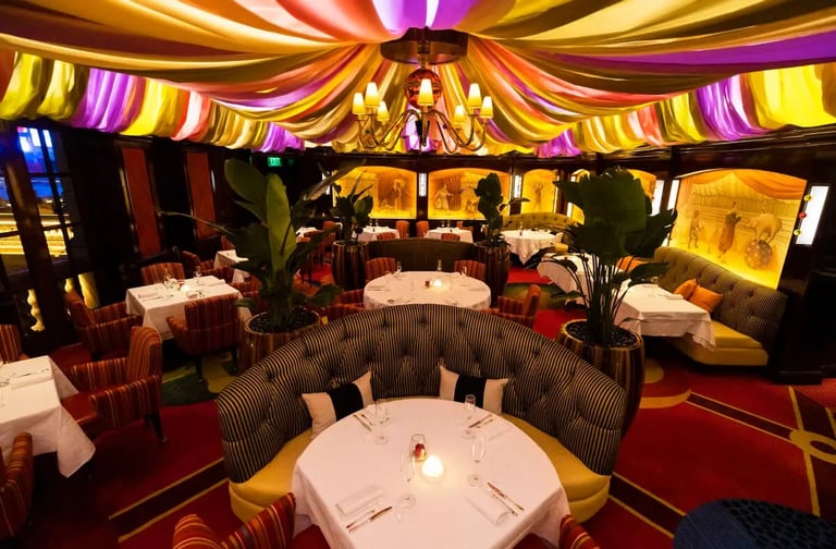 Elegant dining room with colorful striped fabric ceiling draping in yellow, purple, and red, round white tables with place settings, and ambient golden lighting