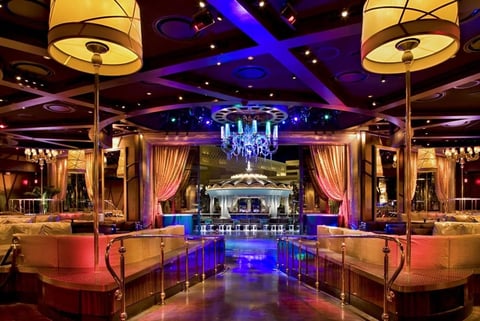 Luxurious nightclub interior with gold lighting, blue neon accents, curved bar seating, and crystal chandelier centerpiece