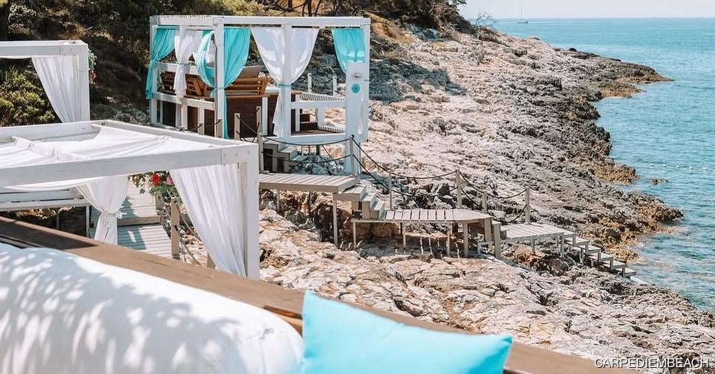 Luxury beach lounge area with white draped cabanas overlooking turquoise Mediterranean waters and rocky coastline