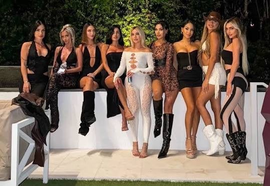 Group of women in dark and neutral party attire posing together at an outdoor event with white furniture and green foliage background