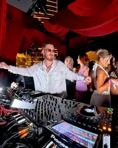 DJ in white shirt performing at nightclub with red lighting and crowd dancing in background