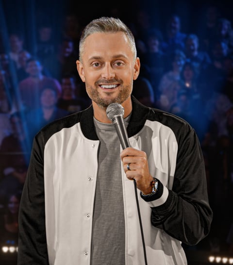 Man smiling while holding microphone on stage, wearing black and white jacket over gray shirt