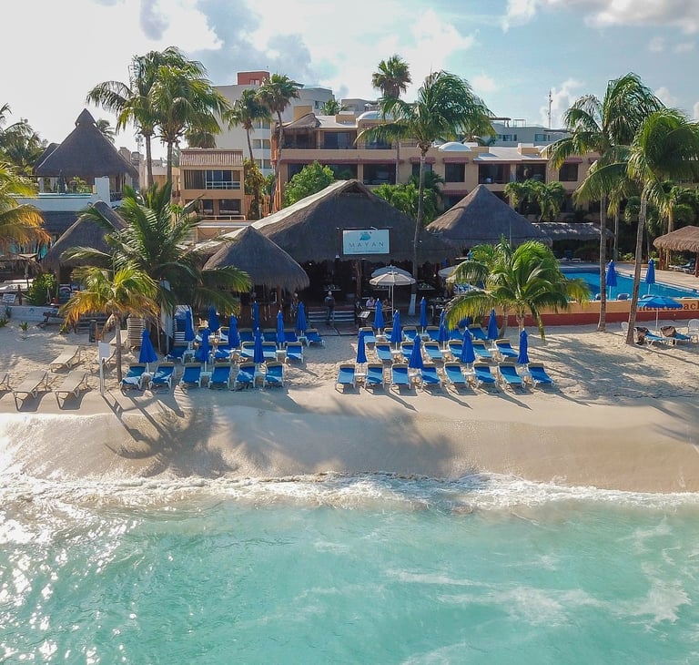 Beach resort with blue umbrellas and lounge chairs on sand, facing turquoise ocean with thatched-roof structures and colorful buildings