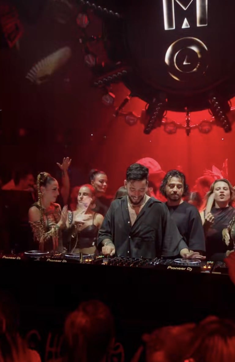 Group of DJs performing at a nightclub with red stage lighting and equipment, audience visible in foreground