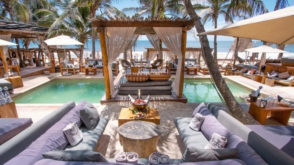 Luxury beach club with turquoise pools, lounge seating, wooden pergolas, palm trees, and ocean views