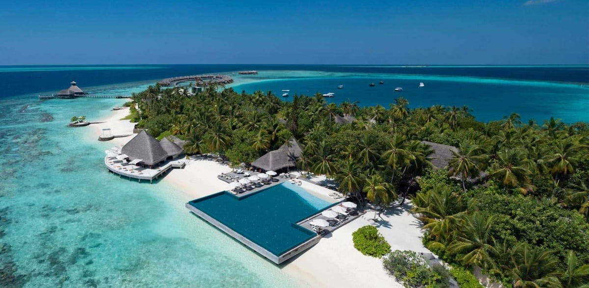 Aerial view of a tropical island resort with overwater bungalows, turquoise lagoon, palm trees, and infinity pool on white sand beach.