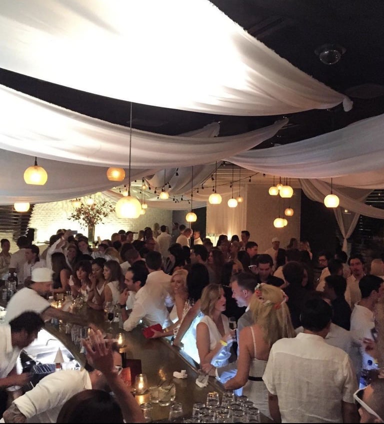 Elegant indoor event with guests in formal attire gathered around illuminated tables under draped ceiling and hanging pendant lights.