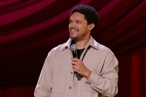Man in beige shirt holding microphone on stage with burgundy curtain background, smiling while performing