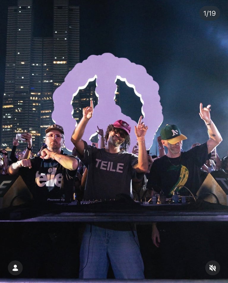 Three DJs perform on stage at night with a purple neon mouth backdrop and city skyline behind them, crowd visible.