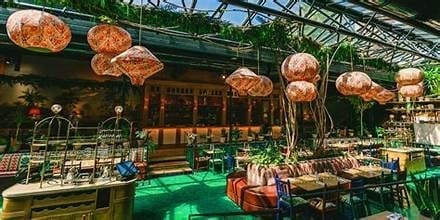 Interior of a modern greenhouse restaurant with hanging copper pendant lights, green neon accents, potted plants, and wooden dining tables