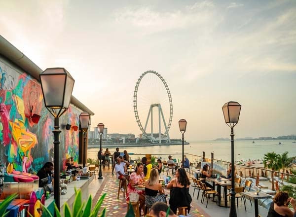 Beachfront promenade with colorful street art, crowds, lamp posts, and Ferris wheel in background at sunset