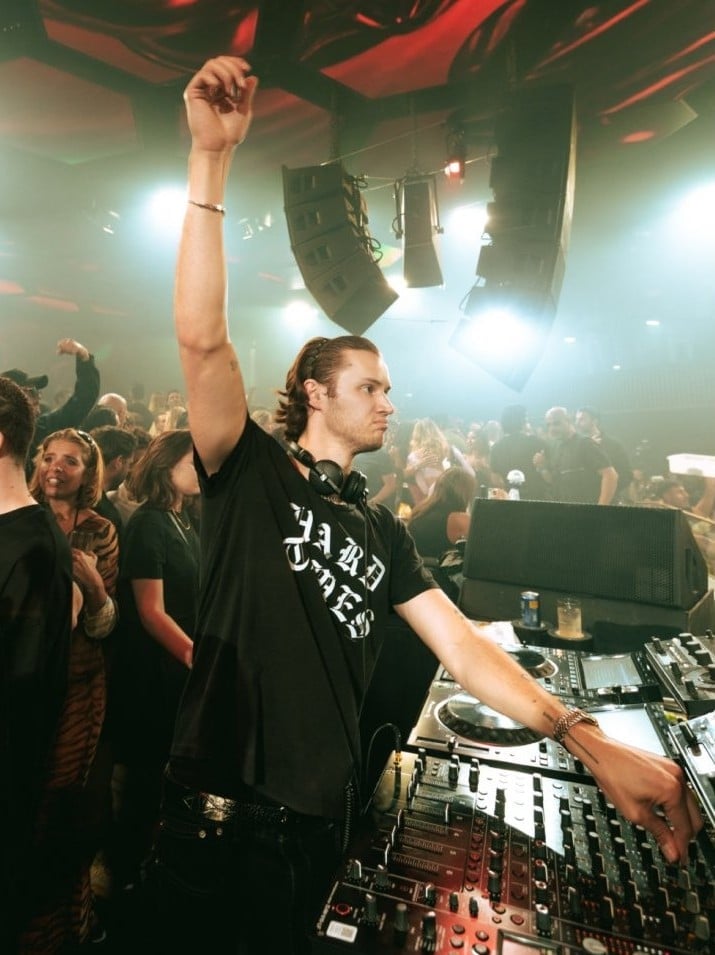 DJ wearing sunglasses and headphones around neck performing at equipment on dark stage with blue lighting