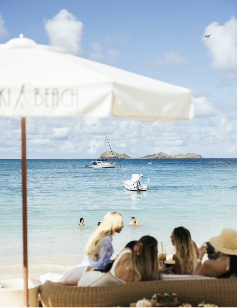 Group of people relaxing on a beach under a white umbrella with turquoise water and sailboats in the background