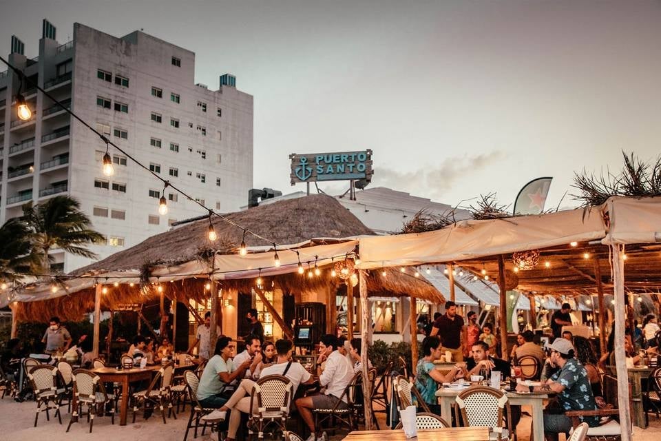 Outdoor restaurant patio with diners seated under warm-lit wooden tent structure at dusk, with city buildings in the background