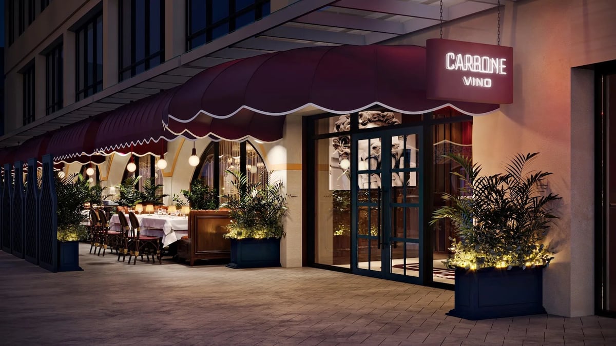 Evening view of Carbone Vino restaurant storefront with warm lighting, dark awning, potted plants, and seating area