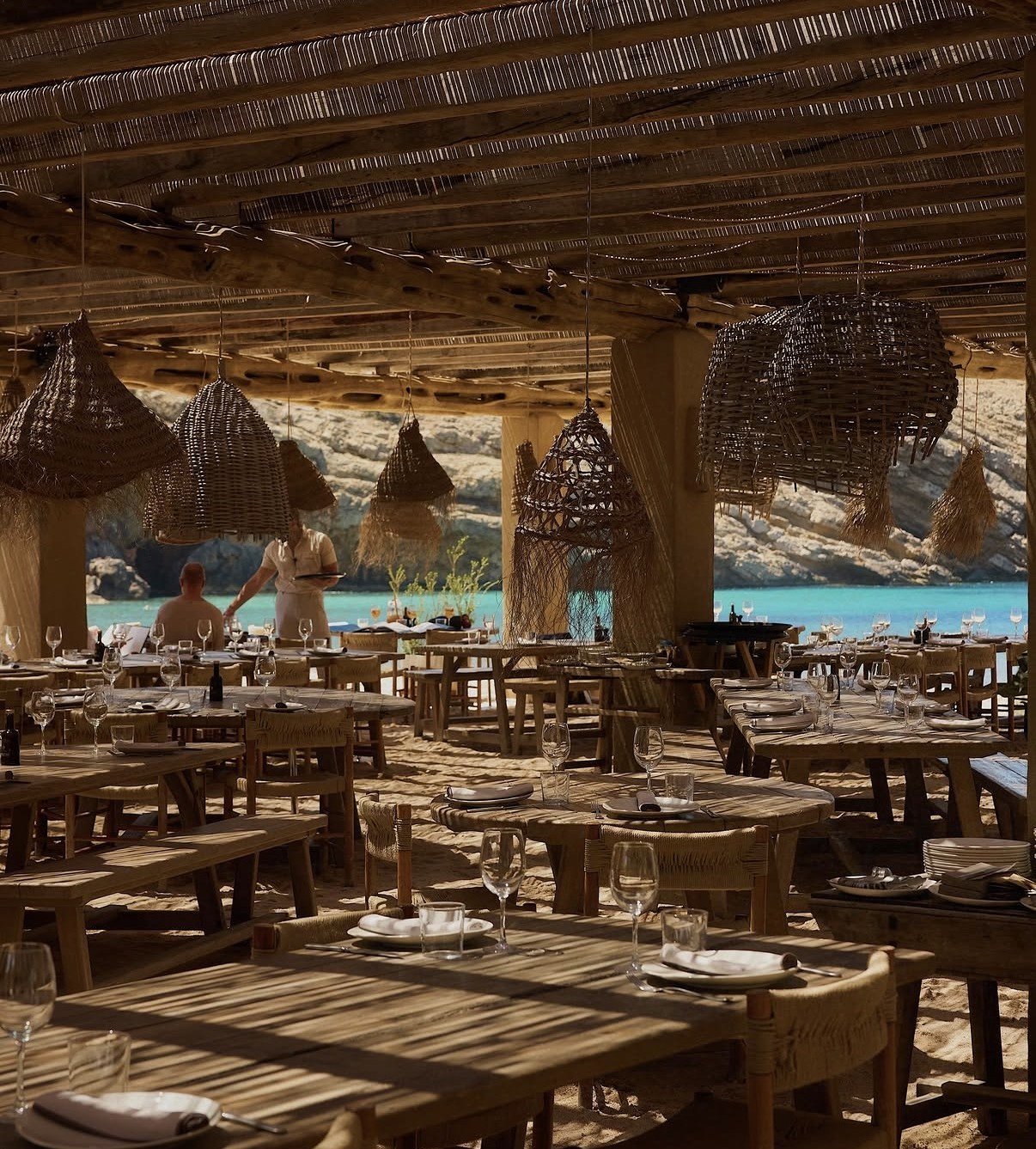 Rustic beachfront restaurant with wooden tables under a thatch roof pergola, overlooking turquoise ocean water
