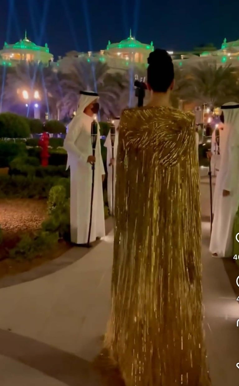 Woman in gold sequined dress walking through illuminated Middle Eastern courtyard at night with ornate buildings and palm trees