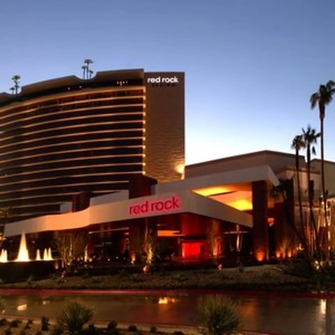 Red Rock Hotel and Casino at dusk with illuminated brown exterior, logo signage, palm trees, and reflection in water