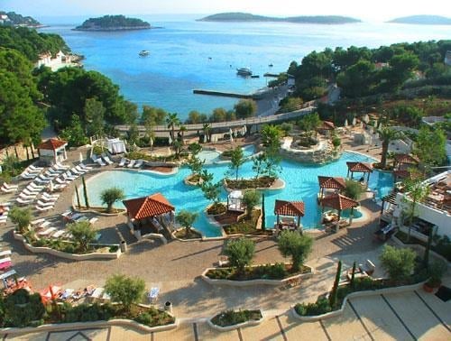 Aerial view of a resort pool complex with turquoise water, loungers, and palm trees overlooking blue islands and sea