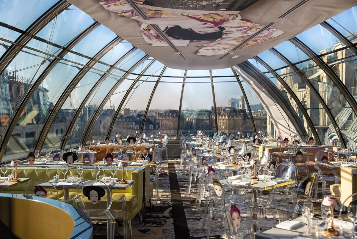 Interior of an upscale glass-domed restaurant with panoramic city views, elegant white table settings, and modern architecture