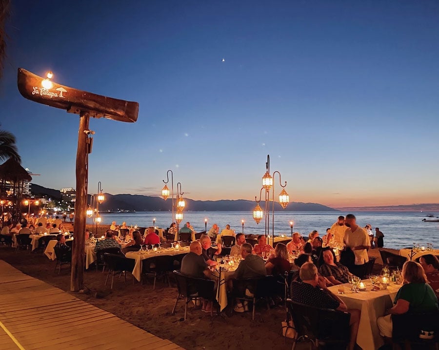 Outdoor beachside dinner party at sunset with guests seated at tables, illuminated by warm lights and lanterns along the shore.