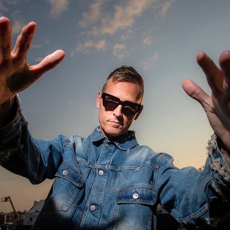 Man wearing sunglasses and denim jacket raising both hands toward camera against cloudy sky